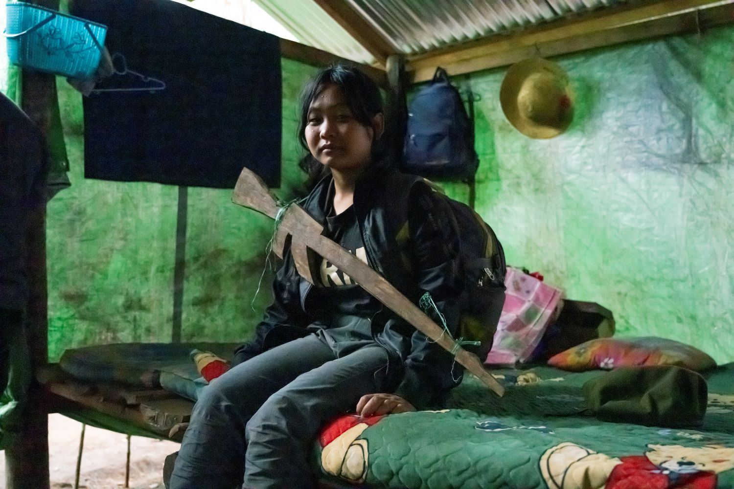 Young woman sits on bed with a fake wooden rifle around her neck