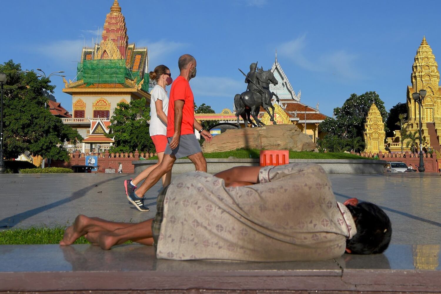 A homeless man lays on the ground as a couple wearing face masks walks past in Phnom Penh, June 23, 2020.