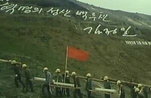 푸른 풀이 난 백두산 암벽에 '혁명의 성산 백두산, 김정일 1992년 2월 16일 새김'이라고 새겨진 김정일 국방위원장의 글발 아래를 천지 돌계단 공사용 돌을 짊어진 노동자들이 지나가고 있다.