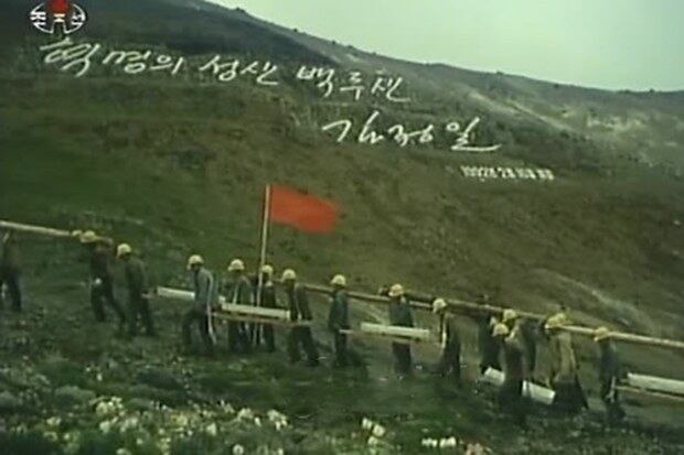 푸른 풀이 난 백두산 암벽에 '혁명의 성산 백두산, 김정일 1992년 2월 16일 새김'이라고 새겨진 김정일 국방위원장의 글발 아래를 천지 돌계단 공사용 돌을 짊어진 노동자들이 지나가고 있다. 
