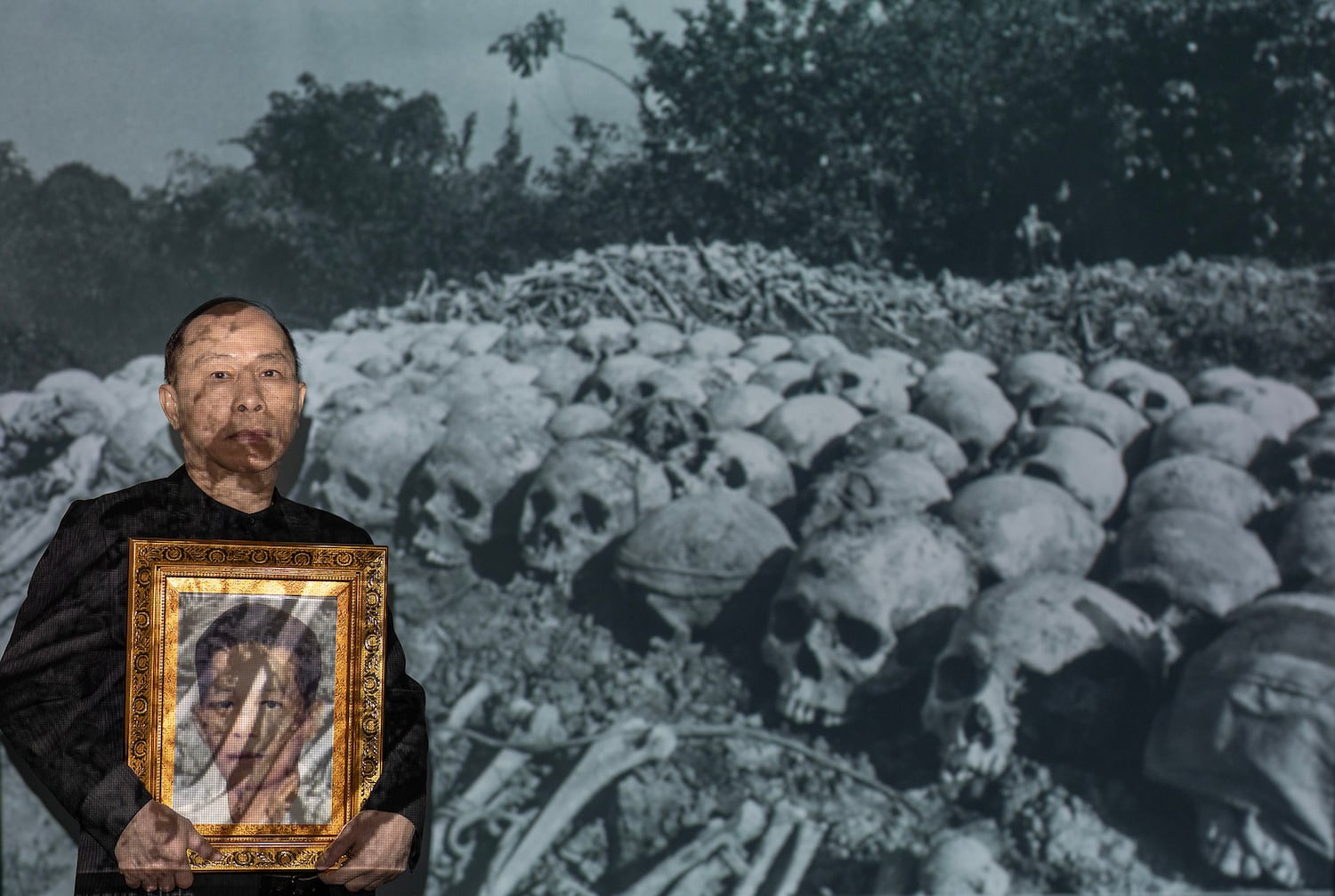 Sum Sok Ry, a senior editor for RFA’s Khmer language service, holds a photo of his father, Sum Bunleng, as he stands in front of a photo of the remains of Khmer Rouge victims.