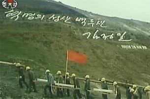 푸른 풀이 난 백두산 암벽에 '혁명의 성산 백두산, 김정일 1992년 2월 16일 새김'이라고 새겨진 김정일 국방위원장의 글발 아래를 천지 돌계단 공사용 돌을 짊어진 노동자들이 지나가고 있다.