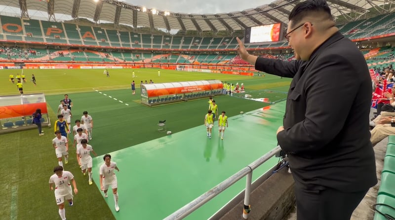 2026 아이치·나고야 아시안게임 남북 여자 축구 조별리그 경기가 끝난 뒤, 김정은 북한 국무위원장 복장을 한 하워드 X 씨가 관중석 난간에서 라커룸으로 퇴장하는 북한 선수들을 향해 손을 흔들고 있다.