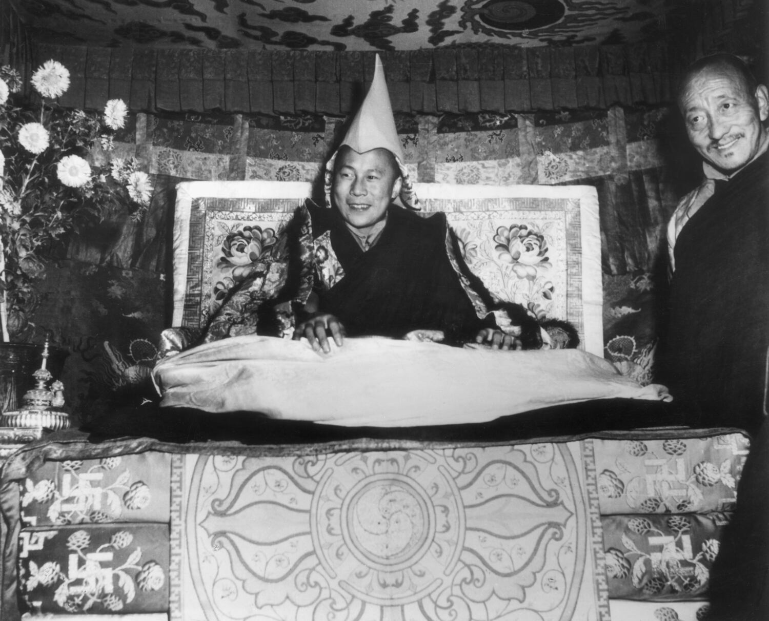 The Dalai Lama, Tenzin Gyatso, seated on his throne and wearing the gold peaked cap which is his Crown, smiles while giving an audience in Lhasa, Tibet in 1959. An assistant monk stands at his side.  (Hulton Archive/Getty Images)